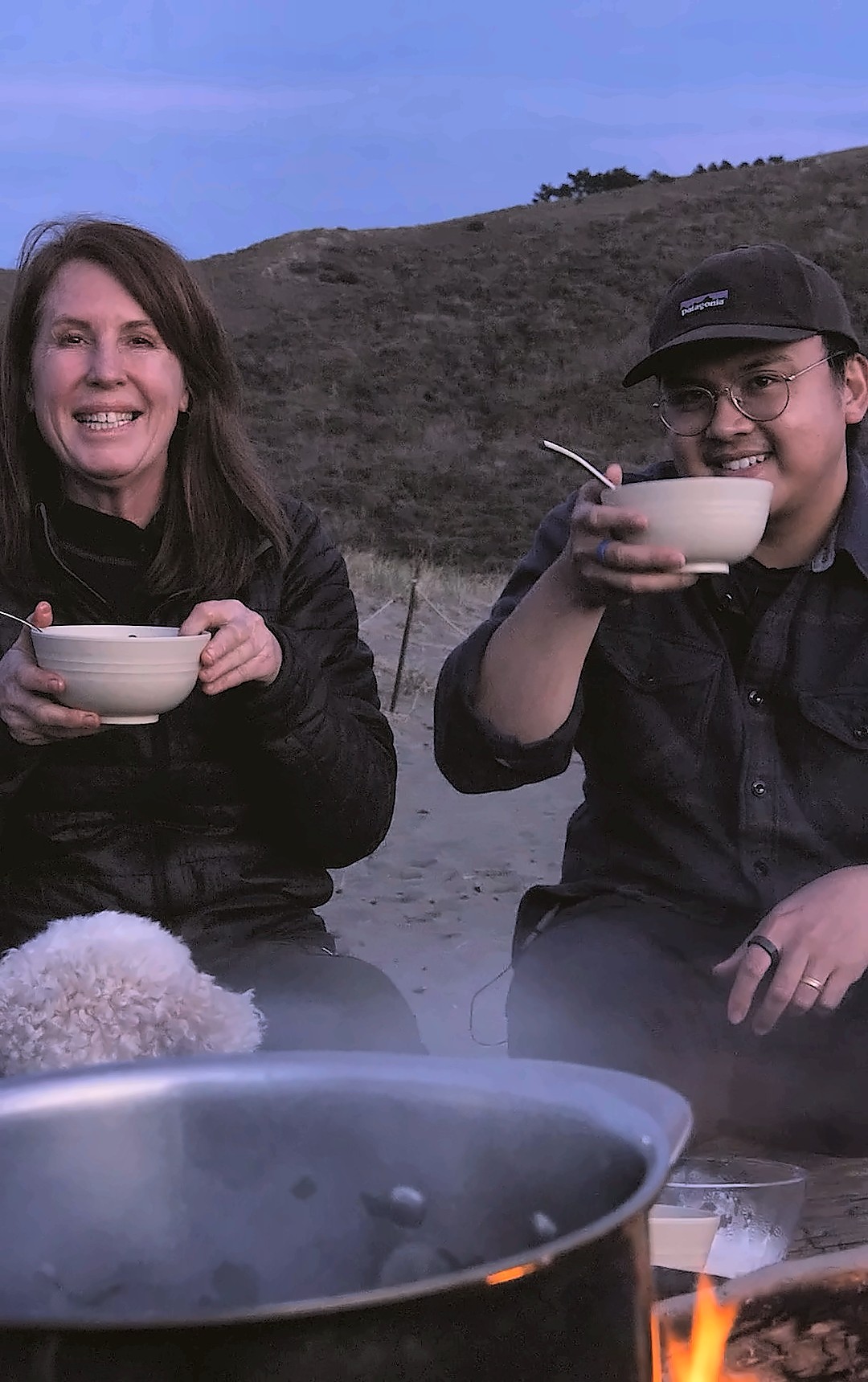 Two people raising bowls of soup around a campfire pot at dusk
