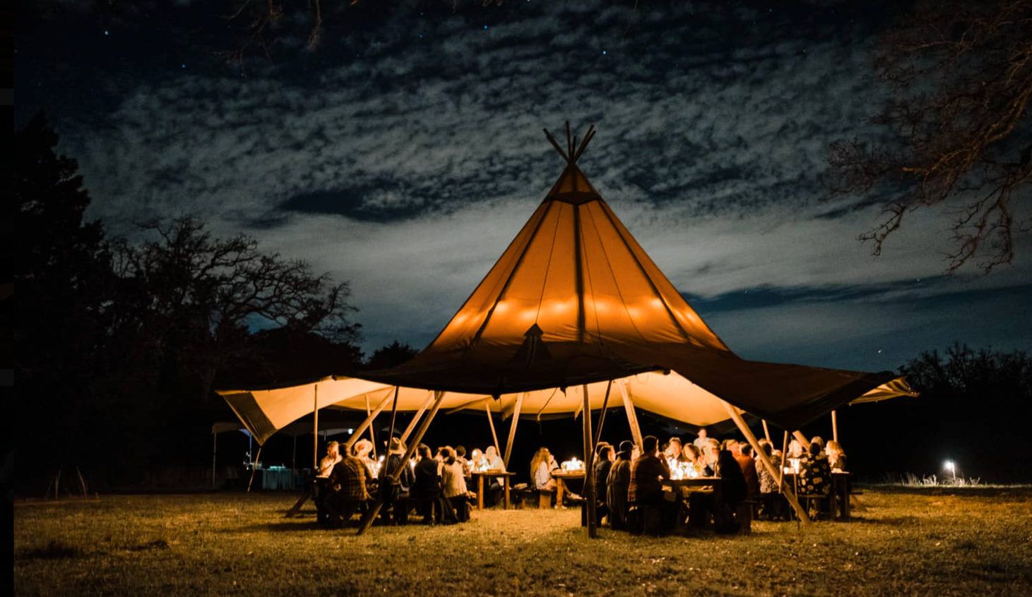A glowing tipi-style tent hosting a long dinner at dusk